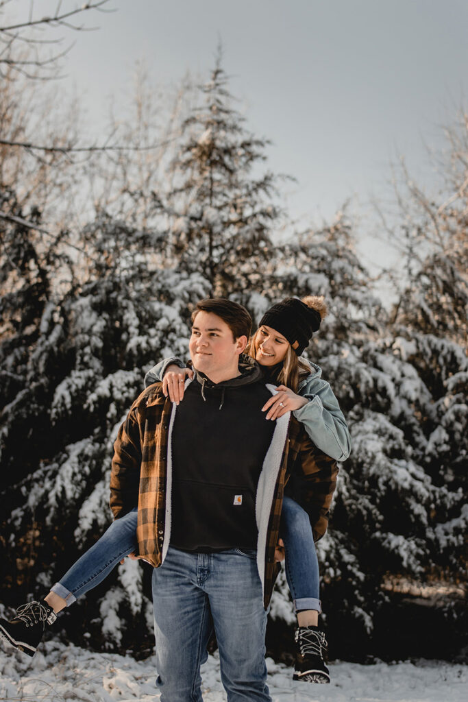 young man looks off smiling as his girlfriend sits piggyback looking his way in a snowy scene 
