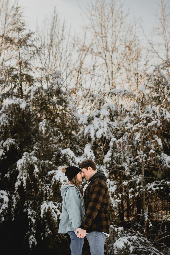 young couple in winter clothes face each other while holding hands in front of snowy evergreens