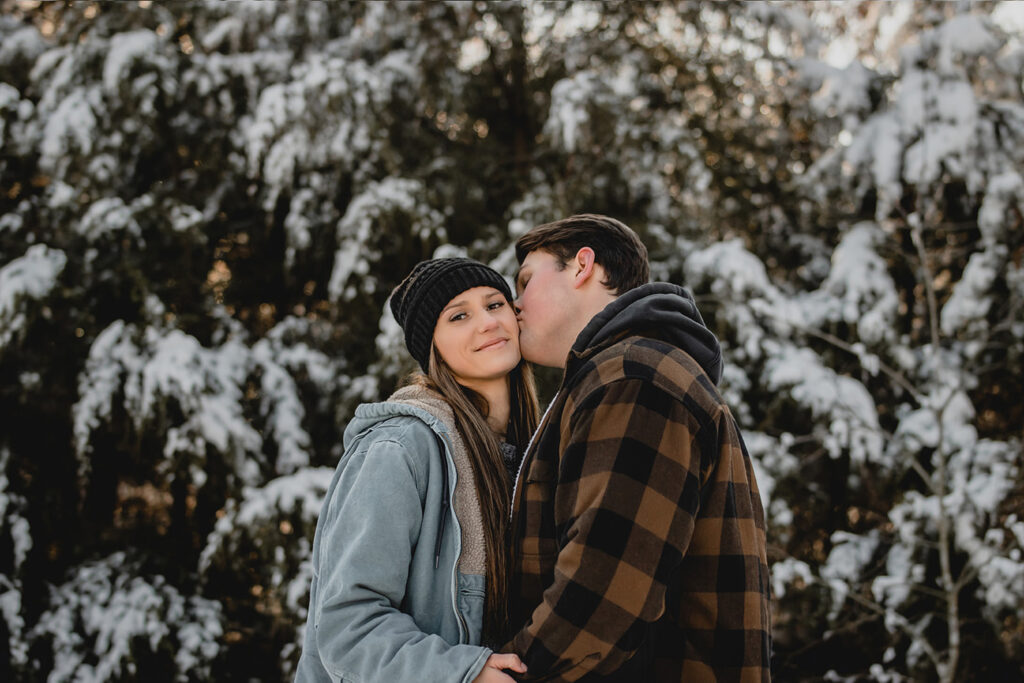 man kissing his girlfriend on the cheek as she smiles to camera in front of snow-covered evergreens