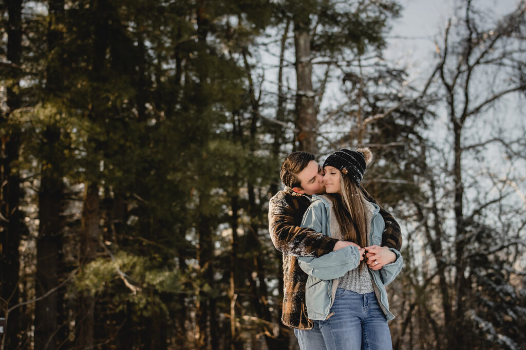 wide shot of man kissing his girlfriend's cheek as she smiles peacefully with her eyes closed in front of a forest of evergreens