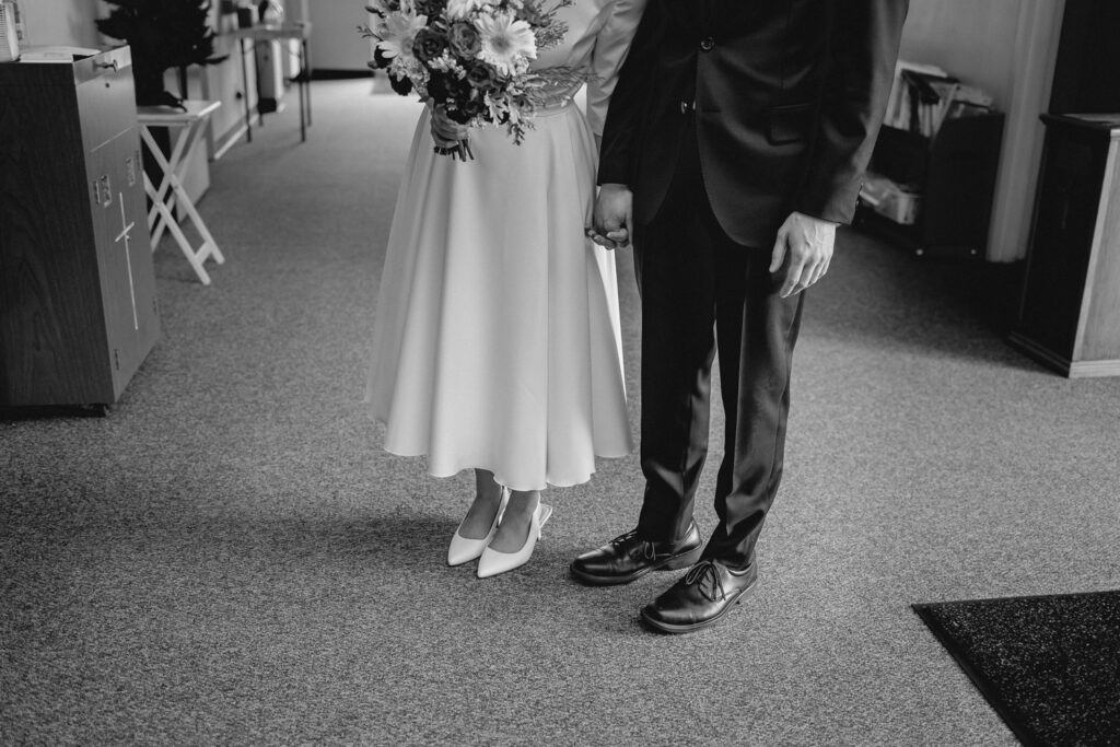 black and white shot from the waist down of bride and groom holding hands in a carpeted entryway