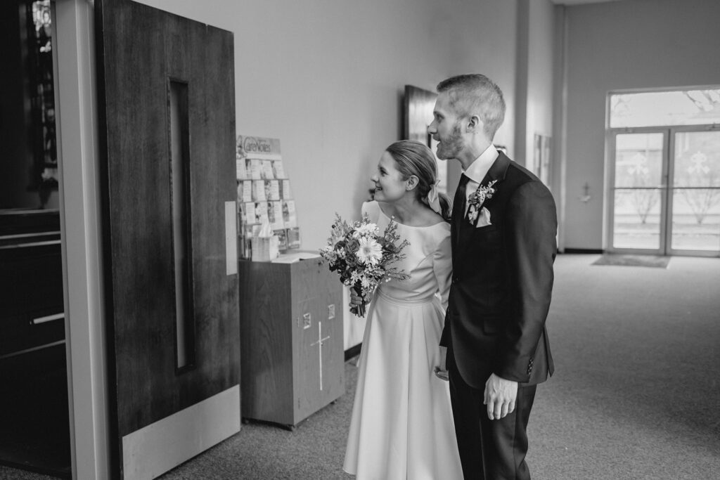black and white shot of bride and groom holding hands and eagerly peeking through the sanctuary doors just before walking down the aisle together 
