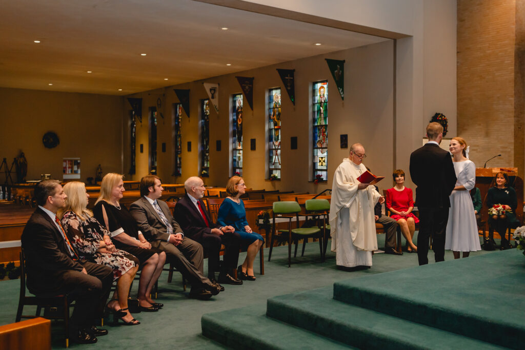 shot from the priest's chair towards the sanctuary pews, a young bride and groom stand before their seated immediate family and priest to be married