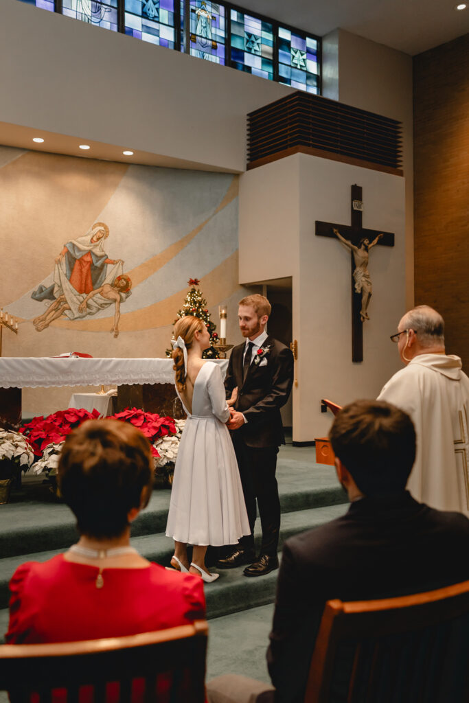 a young bride and groom can be seen through the out of focus silhouettes of the bride's mother and brother as they listen to the priest