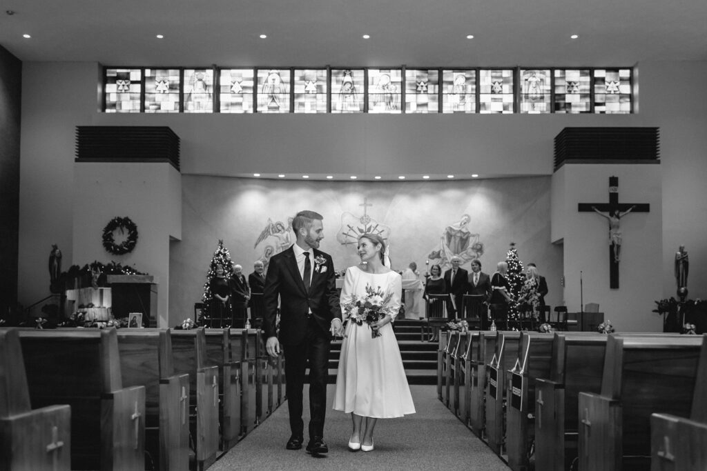 centered shot down the center pew aisle towards the alter and strip of stained glass windows as newlyweds proceed together down the aisle 
