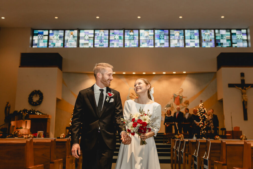 shot down the center pew aisle towards the alter and strip of stained glass windows as newlyweds proceed together down the aisle 
