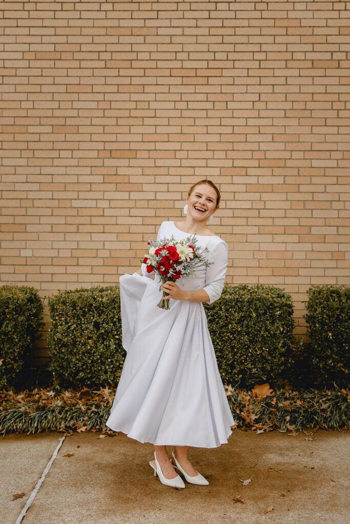 full body portrait of a young bride in simple vintage-inspired white dress holds a bouquet of red flowers and beams as she twirls her skirt in front of a tan brick wall 