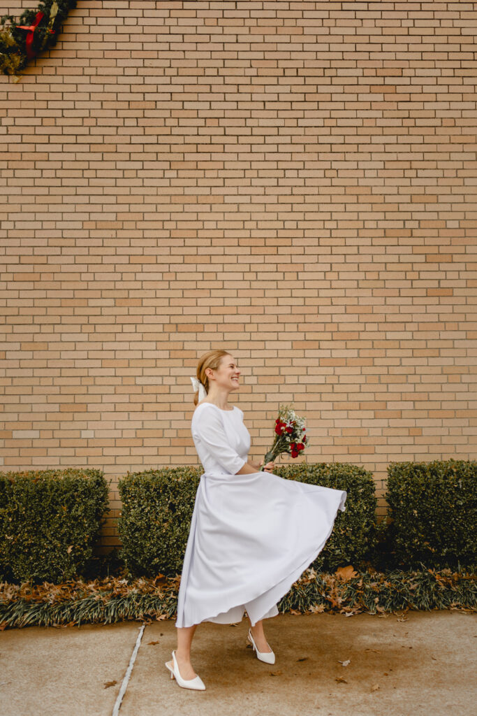 far away shot of a young bride in simple vintage-inspired white dress holds a bouquet of red flowers and beams as she twirls her skirt in front of a tan brick wall 