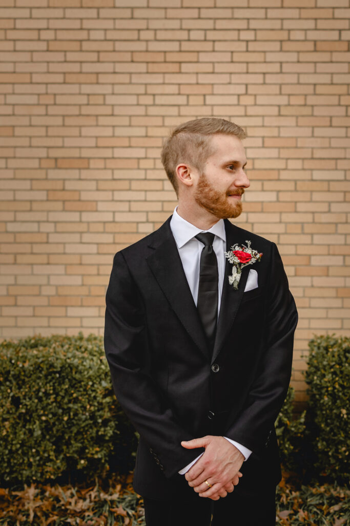 waist up portrait of a young groom in a black tux and tie with red boutonniere looks to his side in front of a tan brick wall 