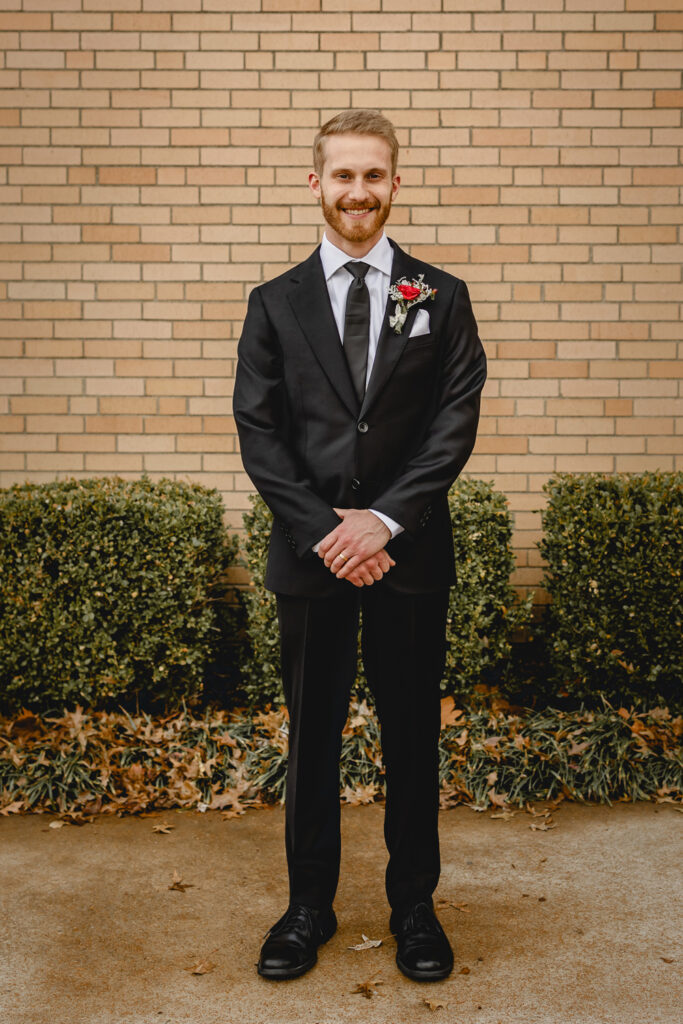 Full bodyportrait of a young groom in a black tux and tie with red boutonniere smiling