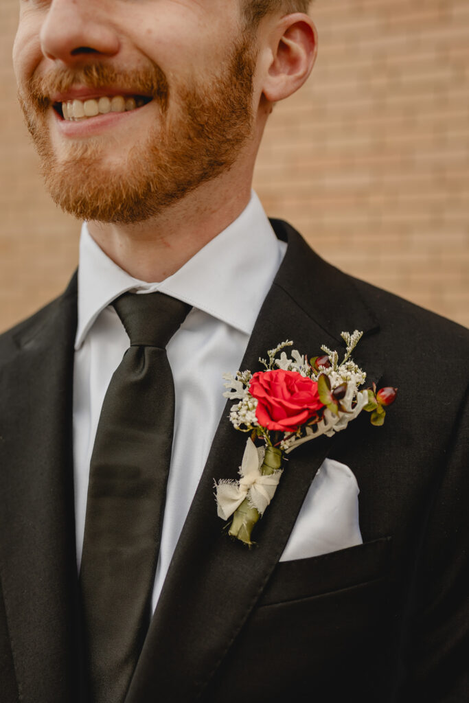 close up detail of a young groom's smile in a black tux and tie and his red boutonniere