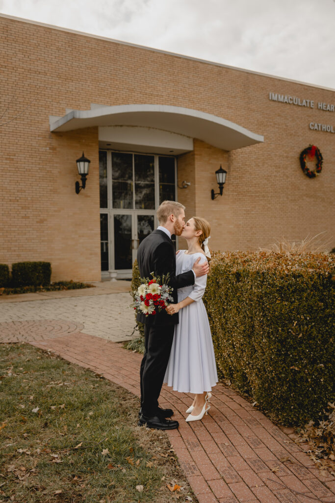 young traditional newlyweds kiss on a brick pathway outside the door of their tan brick church
