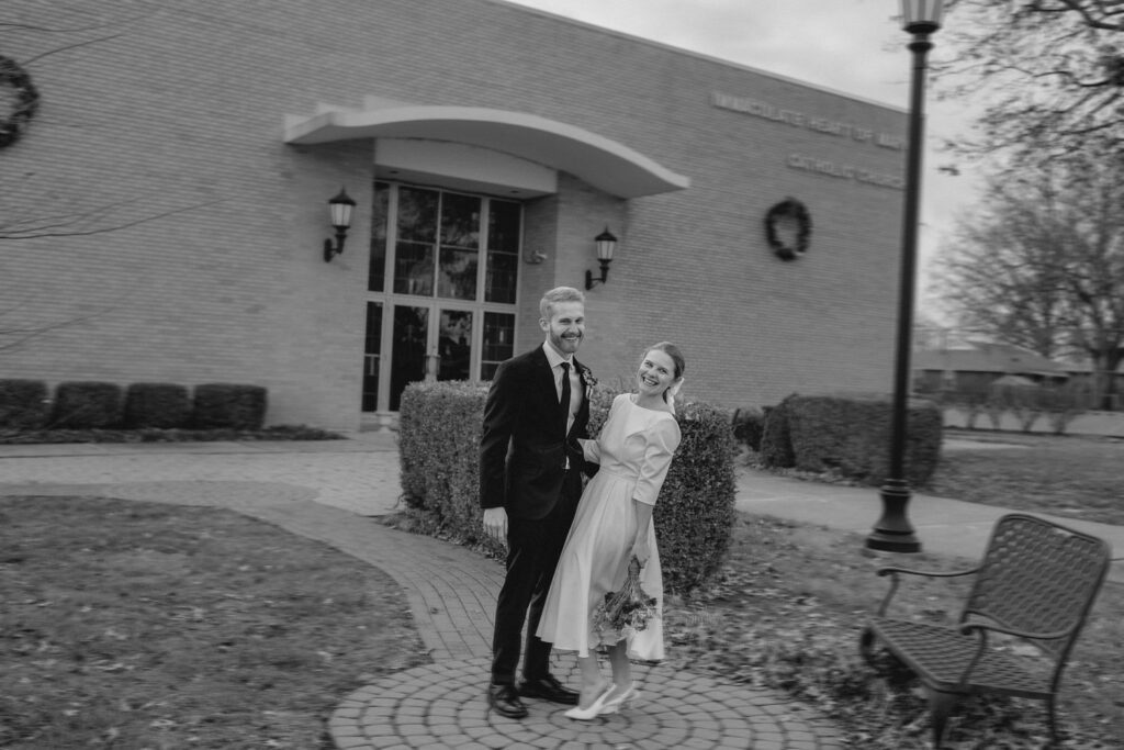 black and white blurry shot of young newlyweds laughing in front of a church in reaction to (off camera) a congratulatory honk from a passerby