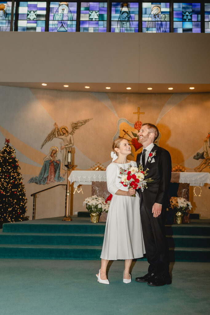 direct-flash colorful shot of bride holding red flowers and smiling at her husband who is laughing atop the teal carpeted church chapel