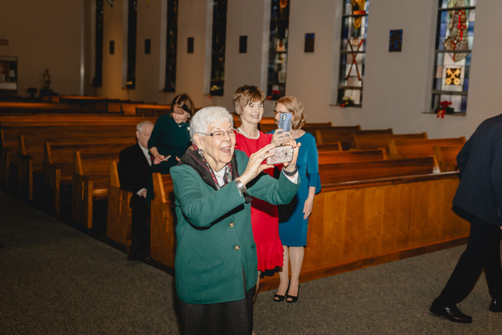 a grandmother excitedly holds up her cell phone to take a picture of the bride and groom