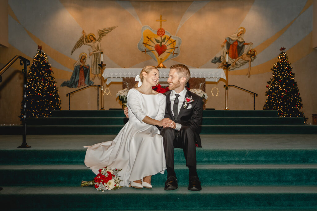 centered direct-flash colorful shot young newlyweds sitting on teal carpeted chapel steps between two lit christmas trees