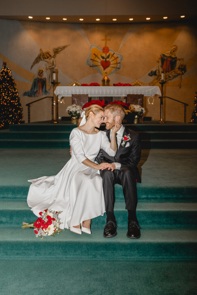 centered direct-flash colorful shot young newlyweds sitting on teal carpeted altar steps and smiling big as they lean their foreheads close together